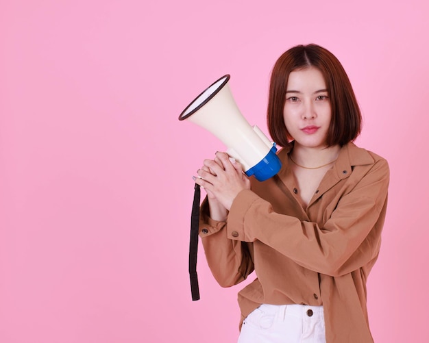 Portrait studio cutout shot of Asian young short hair female model in brown long sleeve shirt standing look at camera smiling holding loudspeaker megaphone shouting announcing news on pink background.