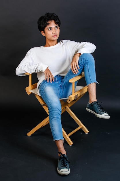 Portrait studio full body shot of Asian young LGBTQ gay glamour handsome bisexual homosexual male model in casual outfit sitting crossed legs on wooden chair posing gesturing on black background