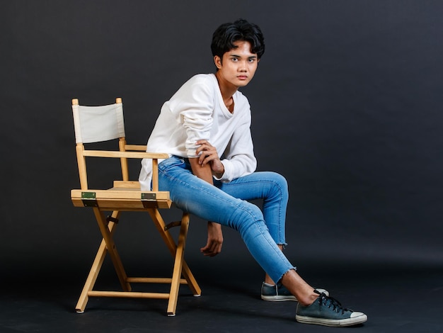 검은 배경에 몸짓으로 포즈를 취하는 나무 의자에 다리를 꼬고 앉아 캐주얼 복장을 한 아시아 젊은 LGBTQ 게이 글래머 잘생긴 양성애자 동성애 남성 모델의 전신 사진