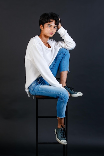 Portrait studio full body shot of Asian young LGBTQ gay glamour handsome male model in casual white shirt jeans sneakers sitting on tall chair laughing holding hand cover mouth on black background