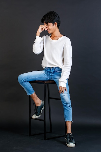 Portrait studio full body shot of Asian young LGBTQ gay glamour handsome male model in casual white shirt jeans sneakers sitting on tall chair laughing holding hand cover mouth on black background