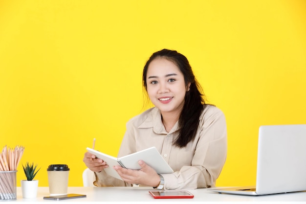 Portrait studio shot of Asian chubby plump female secretary employee worker sitting holding notebook and pencil in hands look at camera smiling at company office working desk on yellow background.