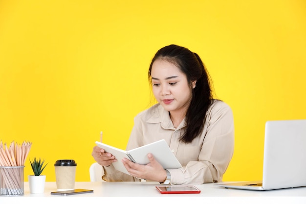 Portrait studio shot of Asian chubby plump female secretary employee worker sitting holding notebook and pencil in hands look at camera smiling at company office working desk on yellow background.
