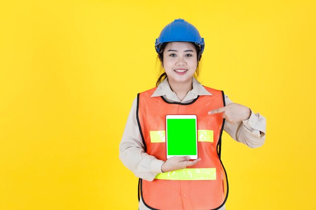 Portrait studio shot Asian female professional engineering foreman manager in hard helmet and reflective safety vest smiling look at camera checking inventory stock from tablet on yellow background.