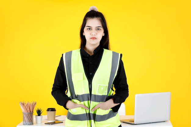 Portrait studio shot of Asian professional confident beautiful female engineer foreman employee wear reflection safety vest outfit sitting on office working desk look at camera on yellow background.