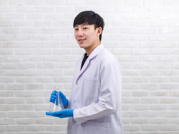 Portrait studio shot Asian professional male scientist in white lab coat rubber gloves standing smiling look at camera holding empty blank Erlenmeyer flask glassware in hands on brick wall background.