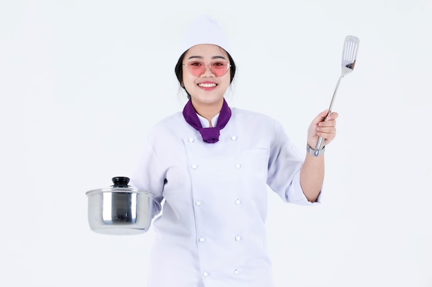白い背景の上のステンレス製の鍋とカバーを保持しているカメラを見て笑顔で立っている料理人の制服とスカーフで女性エグゼクティブシェフを調理するアジアのプロのレストランのポートレートスタジオショット。