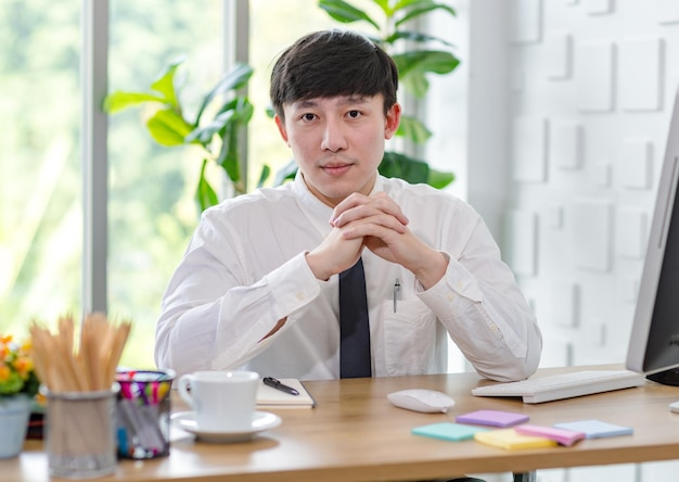 Portrait studio shot of Asian professional successful male businessman employee in formal shirt with necktie sitting look at camera at working desk with computer monitor keyboard mouse and stationery.