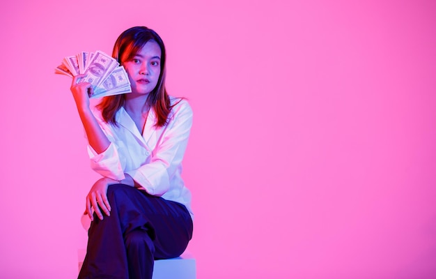 Portrait studio shot of Asian rich wealth female businesswoman model in casual wears sitting look at camera holding full hundred dollars USD cash banknote currency stack on pink light background.