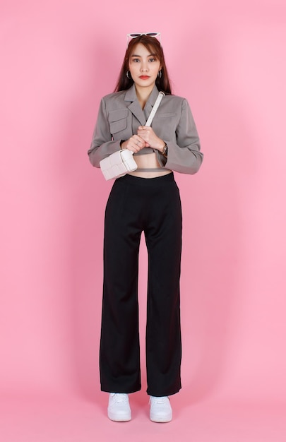 캐주얼 크롭 탑 스트리트에서 유행하는 아시아의 트렌디한 여성 힙스터 10대 모델의 초상화 스튜디오 사진은 분홍색 배경에 카메라를 바라보는 가죽 핸드백 지갑을 들고 재킷 선글라스를 착용합니다.