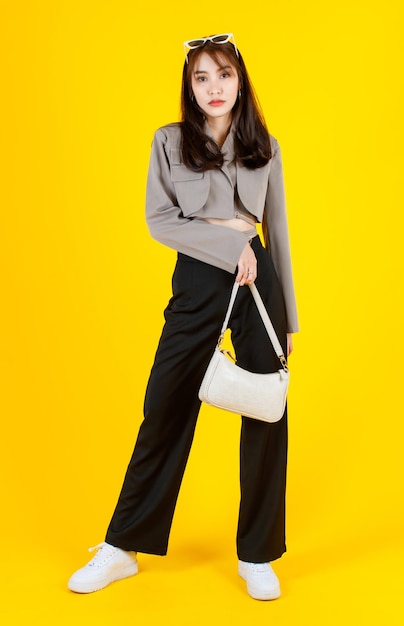 Portrait studio shot of Asian trendy fashionable female hipster teen model in casual crop top street wears jacket sunglasses sneakers holding handbag purse look at camera on yellow background