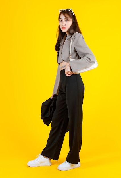 캐주얼 크롭 탑 스트리트에 있는 아시아의 트렌디한 여성 힙스터 10대 모델의 초상화 스튜디오 샷은 핸드백 지갑을 들고 노란색 배경의 카메라를 바라보는 재킷 선글라스 운동화를 착용합니다.