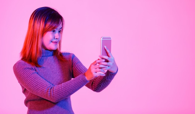 Portrait studio shot of Asian urban female model in gray turtleneck sweater standing smiling look at camera holding touchscreen smartphone in hands chatting with friends on pink light background.