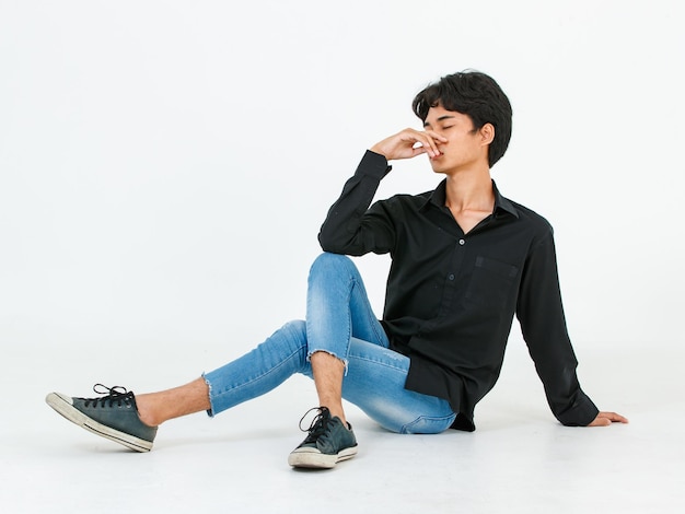 Ritratto in studio di giovane asiatico lgbt gay bisessuale omosessuale glamour maschio modello alla moda in abito casual seduto gambe incrociate sul pavimento occhi chiusi in posa tenendo la mano su sfondo bianco