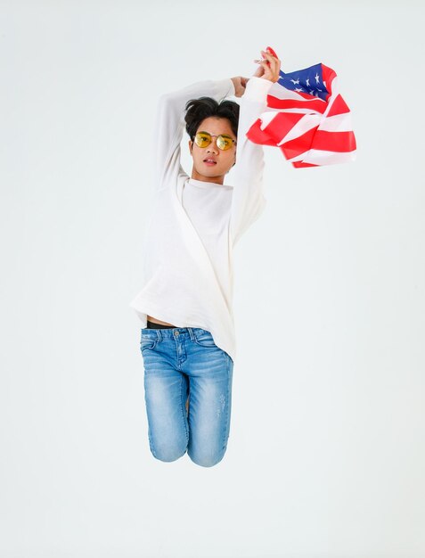 Portrait studio shot of Asian young LGBT gay bisexual homosexual male fashionable model in casual outfit and fashion sunglasses standing holding waving flying USA national flag on white background