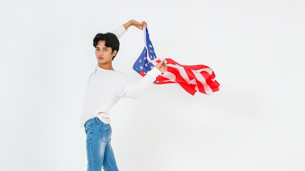 アジアの若いLGBTゲイバイセクシュアル同性愛者の男性のファッショナブルなモデルのカジュアルな服装と白い背景の上の空飛ぶ米国国旗を振って立っているファッションサングラスのポートレートスタジオショット