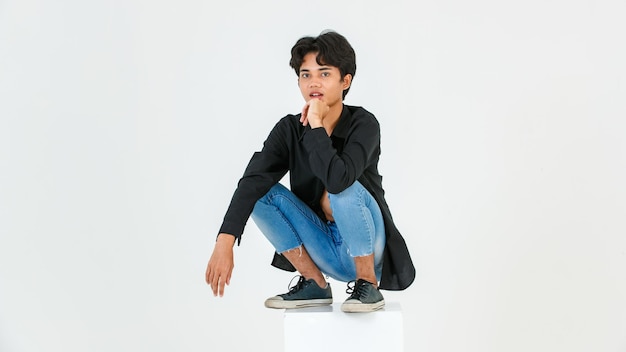 Portrait studio shot of Asian young LGBT gay bisexual homosexual topless male fashionable model in casual black shirt and denim jeans sitting on square boxes posing look at camera on white background