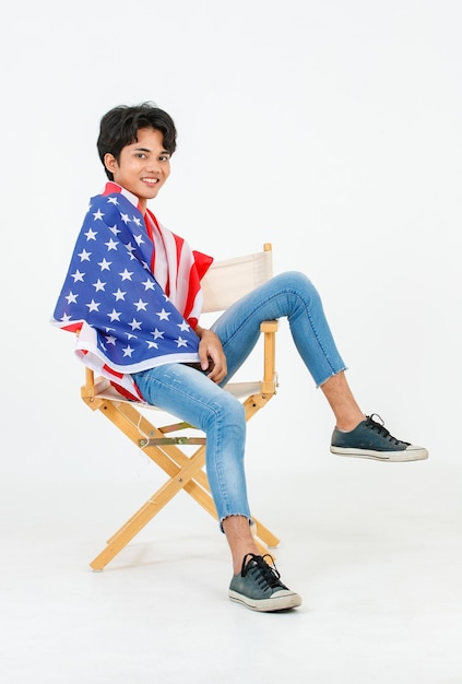 Portrait studio shot of Asian young LGBT gay bisexual homosexual topless male fashionable model sitting on chair smiling using United States of America US national flag cover body on white background