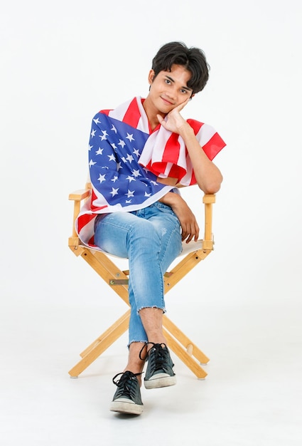 Portrait studio shot of Asian young LGBT gay bisexual homosexual topless male fashionable model sitting on chair smiling using United States of America US national flag cover body on white background