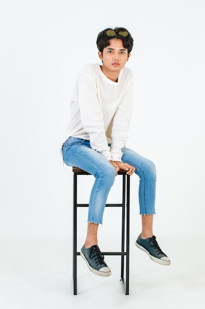 Portrait studio shot of Asian young LGBT gay happy bisexual homosexual male fashionable model in casual outfit fashion sunglasses standing leaning at tall chair smiling posing on white background