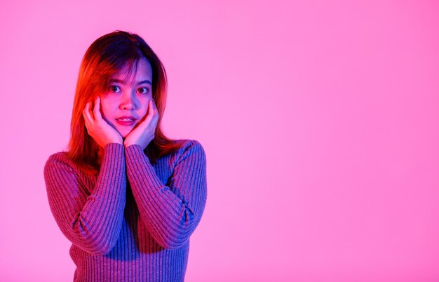 Portrait studio shot of Asian young teen beautiful shy dyed hair female model in long sleeve turtleneck sweater shirt standing posing holding hands on cheeks look at camera on pink light background.