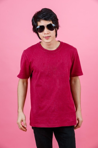 Portrait studio shot of Asian young urban teenager fashion male model in casual street style outfit t shirt and black jeans with aviator sunglasses standing posing look at camera on pink background