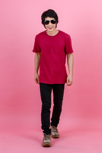 Portrait studio shot of Asian young urban teenager fashion male model in casual street style outfit t shirt and black jeans with aviator sunglasses standing posing look at camera on pink background