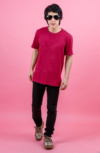 Portrait studio shot of Asian young urban teenager fashion male model in casual street style outfit t shirt and black jeans with aviator sunglasses standing posing look at camera on pink background