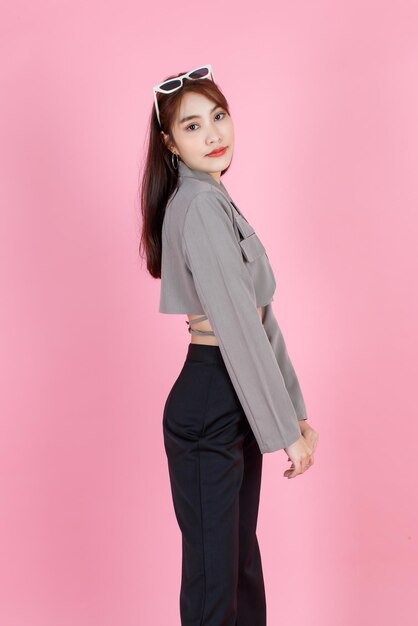 Portrait studio shot of Asian young urban trendy female hipster teenager fashion model in casual crop top street wears jacket holding sunglasses on head standing look at camera on pink background.