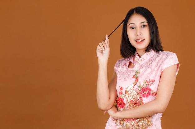 Portrait studio shot of millennial Asian female model in pink cheongsam qipao traditional festival peacock &amp; flowers pattern dress playing rolling hair standing smiling posing on brown background.