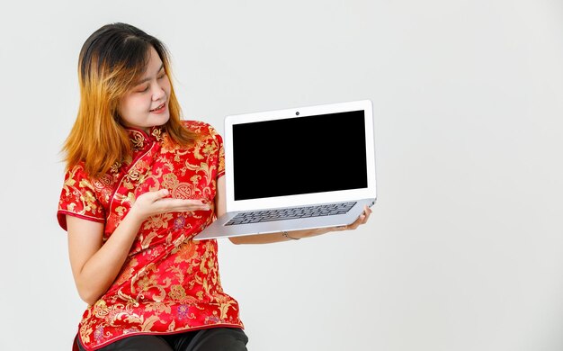 Portrait studio shot millennial Asian female model in red Chinese traditional cheongsam qipao shirt sitting holding showing presenting black blank screen laptop computer product on white background.