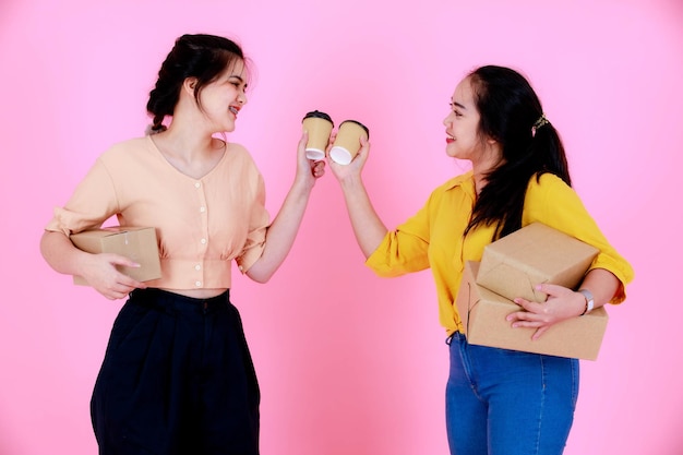 Portrait studio shot of two Asian chubby and slim braces teeth ponytail female business partner standing smiling cheers coffee cup and holding delivery package shipment together on pink background.