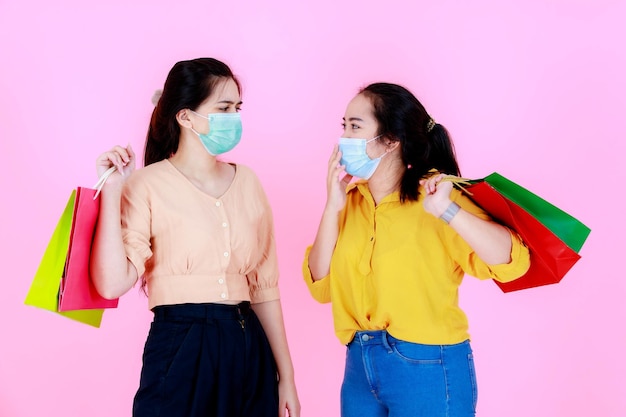 Portrait studio shot of two Asian young happy female shoppers friend wearing face mask and casual outfit carrying colorful shopping bags together during covid pandemic quarantine on pink background.