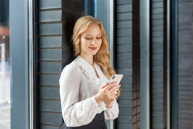 Портрет потрясающей элегантной деловой женщины с длинными светлыми волосами в белой стильной рубашке, опирающейся на кирпичную стену и счастливо улыбающейся, используя смартфон, читающий сообщение в социальной сети в помещении