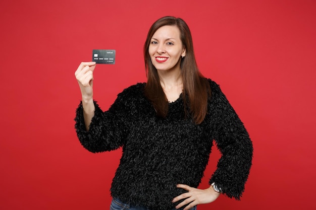 Portrait of stunning smiling young woman in black fur sweater holding credit bank card isolated on bright red wall background in studio. People sincere emotions, lifestyle concept. Mock up copy space.