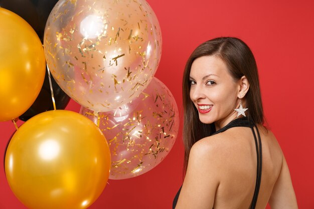 Portrait of Stunning young woman in black clothes celebrating, looking back isolated on bright red background air balloons. St. Valentine's Day, Happy New Year, birthday mockup holiday party concept.