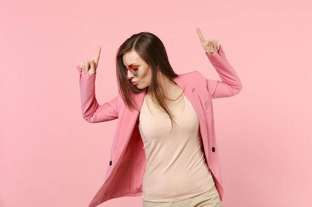 Portrait of stunning young woman in heart glasses dancing, pointing index fingers up isolated on pastel pink wall background in studio. People sincere emotions, lifestyle concept. Mock up copy space.