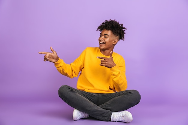 Portrait of stylish african american guy smiling and pointing fingers aside at copyspace while sitting on floor with legs crossed, isolated over violet background