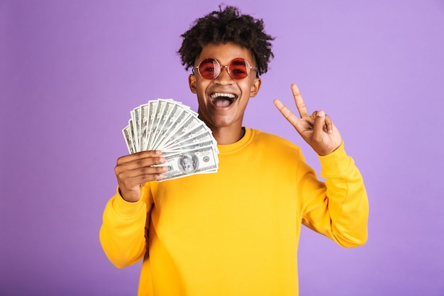 Portrait of stylish african american man wearing sunglasses smiling while holding dollars money banknotes, isolated over violet background