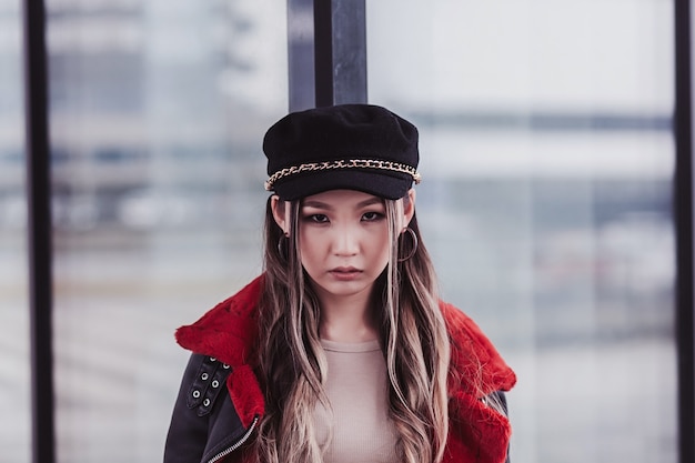 Portrait stylish asian girl wearing hat and jacket with red fur posing in city street and looking at camera