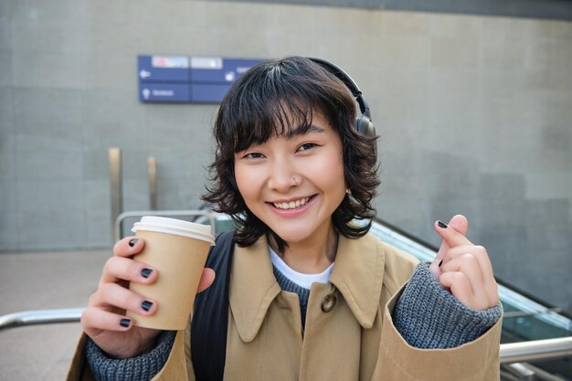 헤드폰을 끼고 있는 세련된 아시아 여성의 초상화는 커피를 마시고 미소는 카푸치노를 즐기는 동안