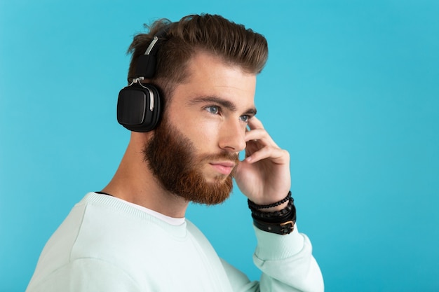 Портрет стильного привлекательного молодого бородатого мужчины, слушающего музыку на беспроводных наушниках в современном стиле, уверенное настроение, изолированное на синем фоне