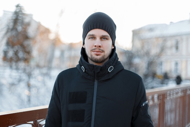 Photo portrait of a stylish attractive young man with a beard in a stylish winter black jacket in a knitted hat on a warm winter day. brutal fashionable guy.