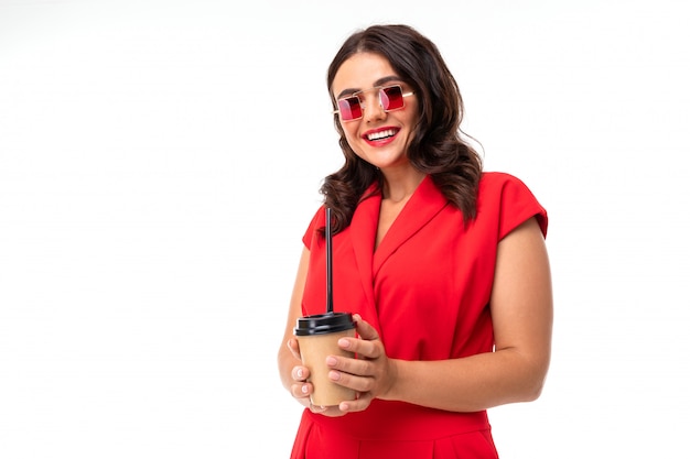 Portrait of stylish brunette girl in sunglasses and a red dress with a glass on white