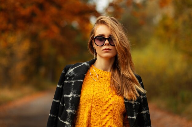 Portrait of a stylish caucasian girl with neutral emotions with fashionable sunglasses in an autumn yellow sweater and black coat walks in a park with orange autumn foliage, outdoors