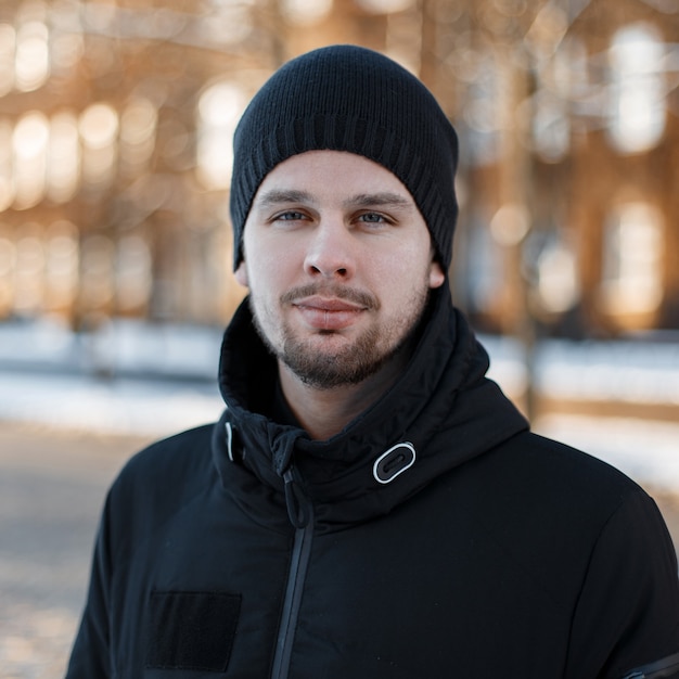 Portrait of a stylish friendly handsome young man with a beard in a fashionable winter black jacket in a black knitted hat on  vintage modern buildings. Attractive fashionable guy.