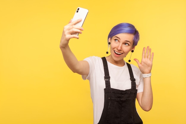 Portrait of stylish friendly hipster girl with violet short hair in denim overalls holding cell phone making video call waving hand to say hello hi online conversation yellow background studio shot