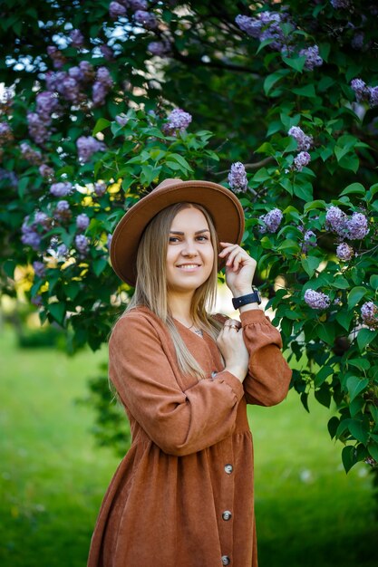 Portrait of a stylish girl in a brown hat and dress on a lilac background. Young woman of European appearance with a smile on her face