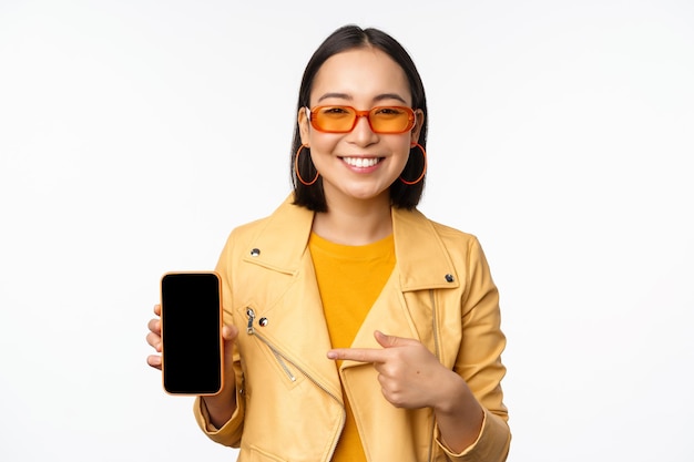 Portrait of stylish korean girl in sunglasses smiling pointing finger at smartphone screen showing mobile phone application standing over white background