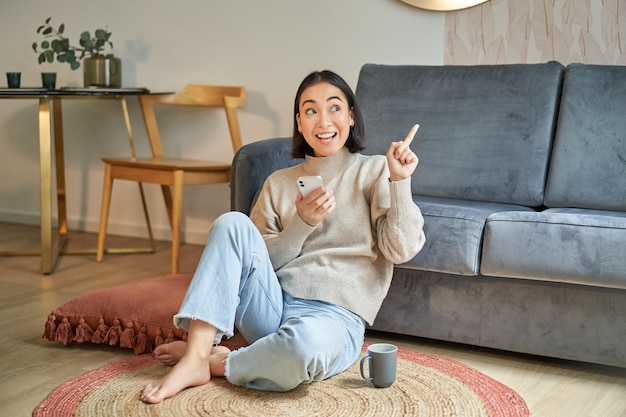 portrait of stylish korean woman sits on floor with smartphone using mobile phone smiling pleased co